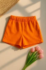 Canlı portakal renkli kadın keten şort, düz zeminde pembe lale çiçekleri ve örgü dokulu hasır parça eşliğinde sergilenmiş.