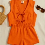 Canlı portakal tonlarında kadın keten alt-üst takım; yelek ve şort kombinasyonu, hasır çanta ve güneş gözlüğüyle düz zeminde sergilenmiş.