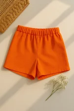 Canlı turuncu renkte kadın keten şort, düz bej zemin üzerinde hasır kutu ve beyaz kuru çiçekle birlikte sergilenmiş.