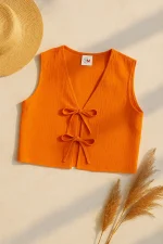 Turuncu keten kadın yeleği, iki fiyonk detayıyla düz bir zeminde, yanında hasır şapka ve kuru pampas otlarıyla birlikte sergilenmiş.